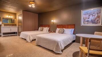 Quarto de hotel com duas camas, banheiro com toucador, uma mesa pequena e um quadro na parede.