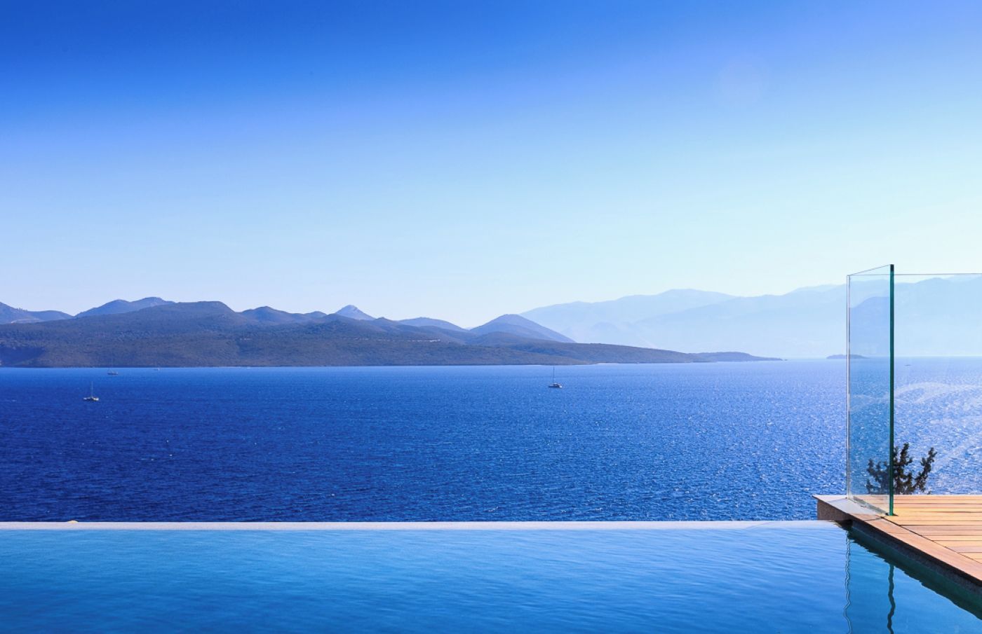 A clear view of a large body of water with mountains in the distance, a wooden deck, and a glass barrier.