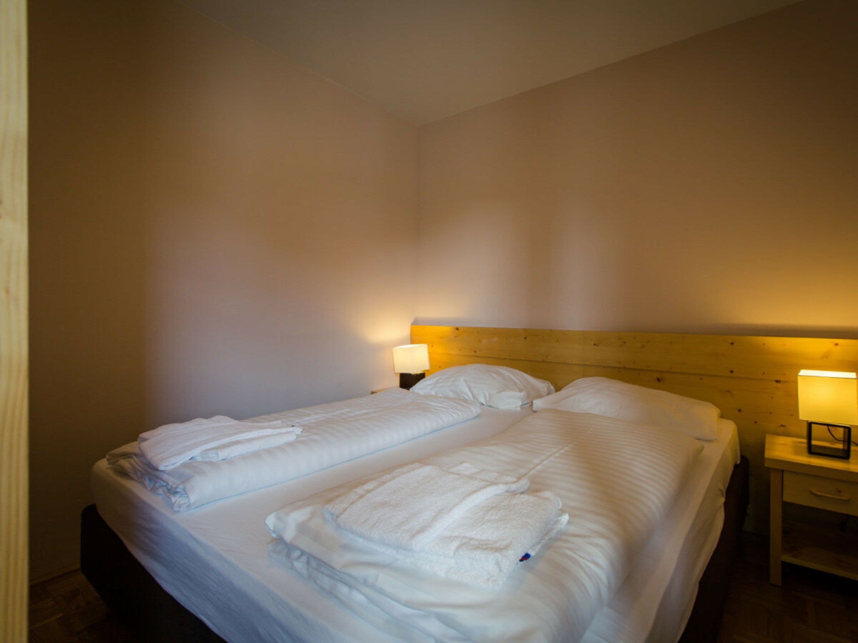 A double bed with two pillows and a folded towel, a wooden headboard, and two bedside lamps.