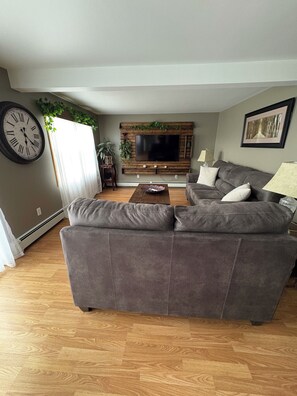 Un salon avec un canapé gris, une table basse en bois, un téléviseur à écran plat fixé à un mur en bois, une horloge et une plante.