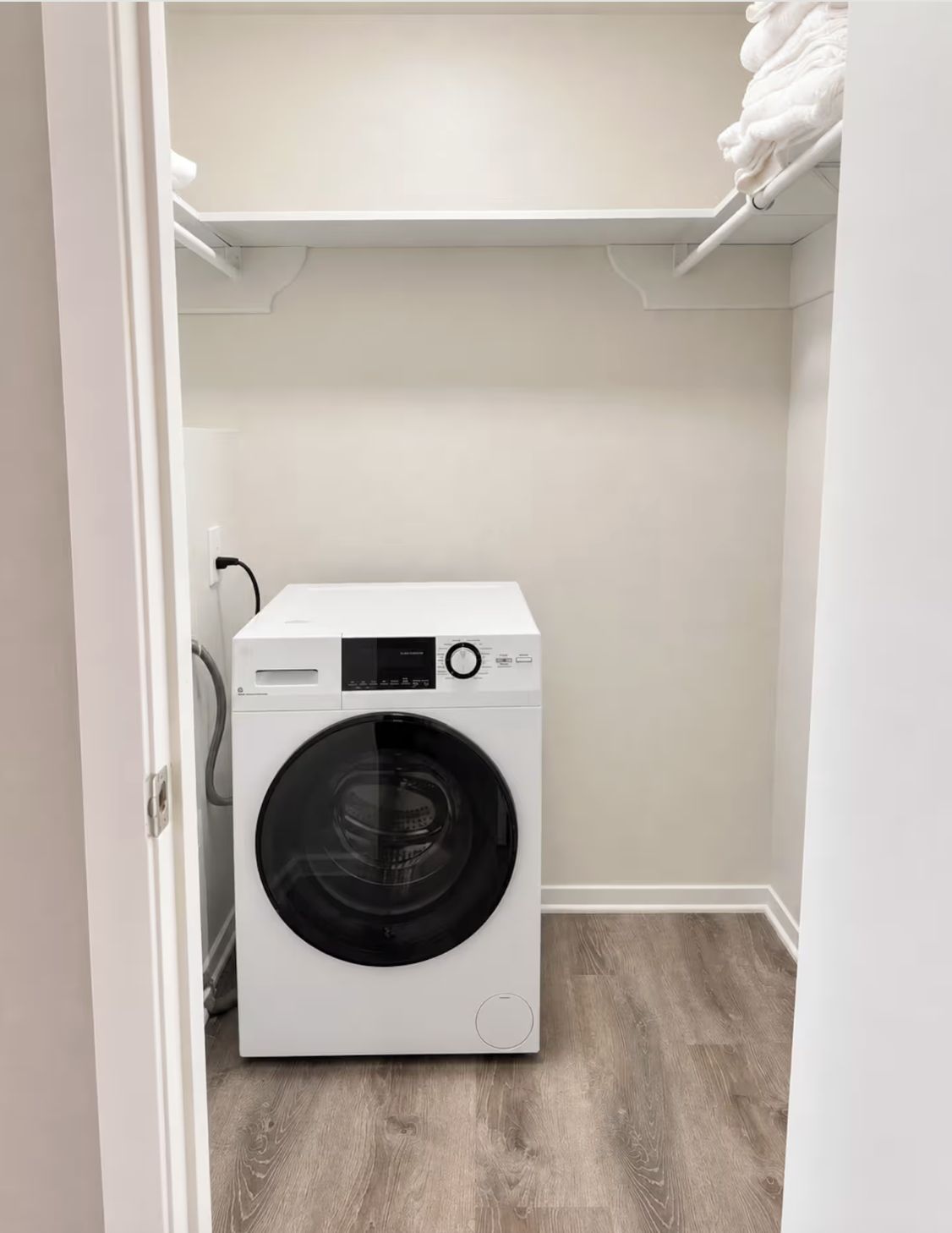 Een compacte, witte wasmachine met frontlader in een kleine wasruimte met een houten vloer en witte muren.