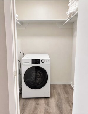 A compact, white front-loading washing machine in a small laundry room with a wooden floor and white walls. - Modern 2BR Near NASA: Washer/dryer, Pool, Balcony (Houston)