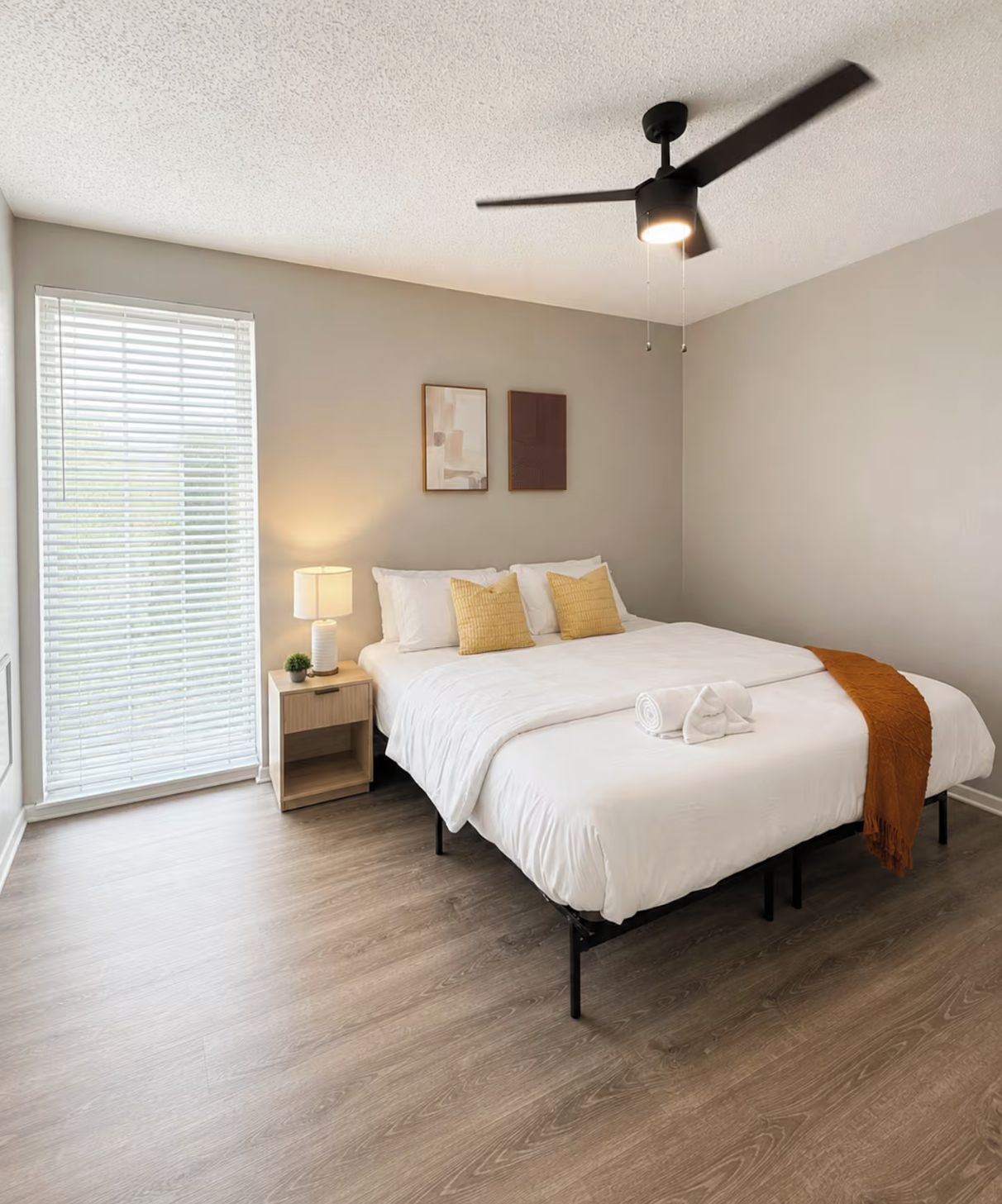 A bedroom with a bed, a nightstand, a lamp, a ceiling fan, and a window with blinds.