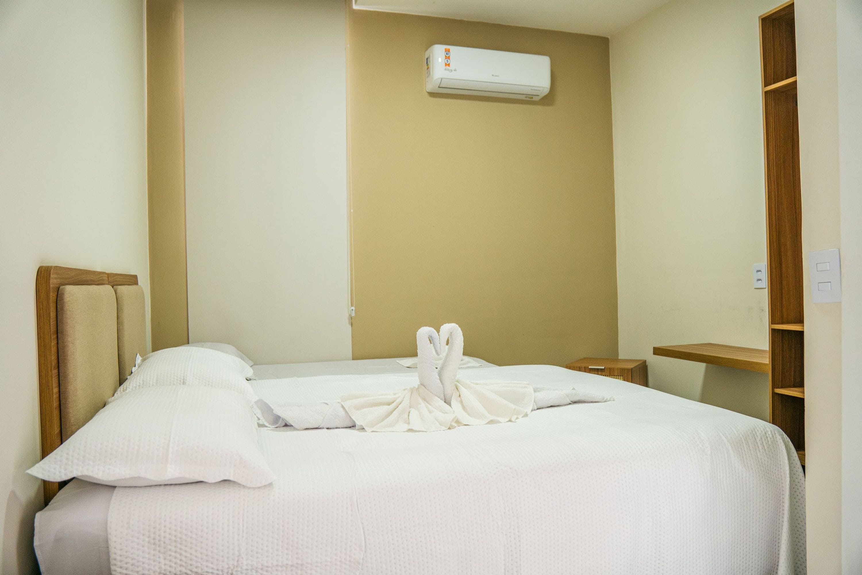 A hotel room with a bed, a wooden headboard, a bedside table, a nightstand, a wardrobe, and an air conditioning unit on the wall.