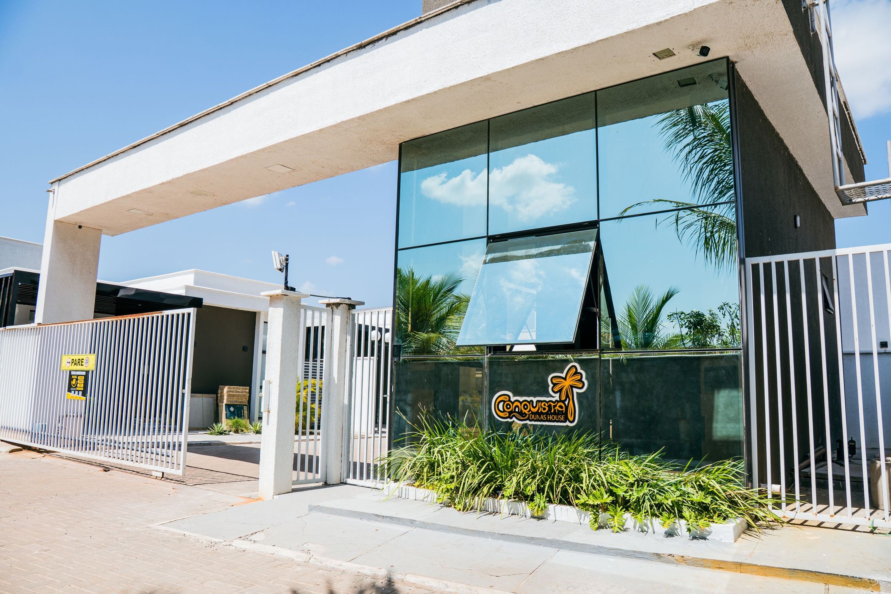 Un edificio moderno con fachada de vidrio, un cartel que dice 'Conquista Dunas House' y una entrada con portón.