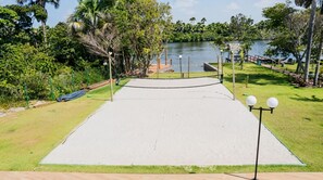 A concrete tennis court with a net, surrounded by grass and trees, with a body of water in the background.