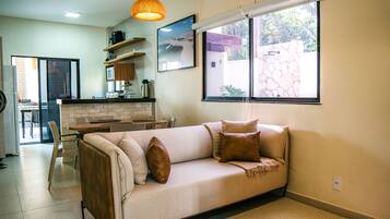 A living room with a sofa, a coffee table, and a dining area with chairs. There is a kitchen visible in the background.