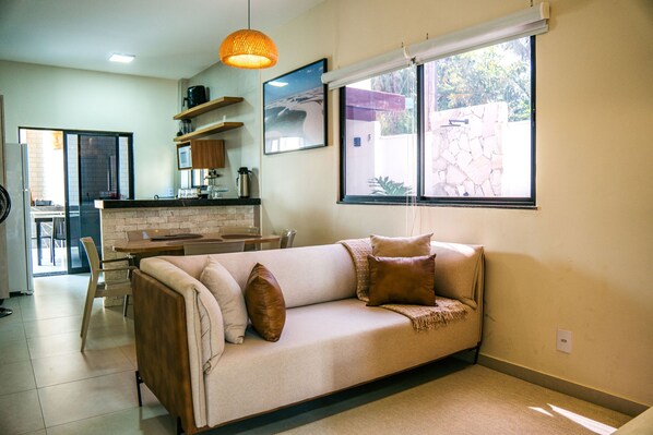 A living room with a sofa, a coffee table, and a dining area with chairs. There is a kitchen visible in the background.