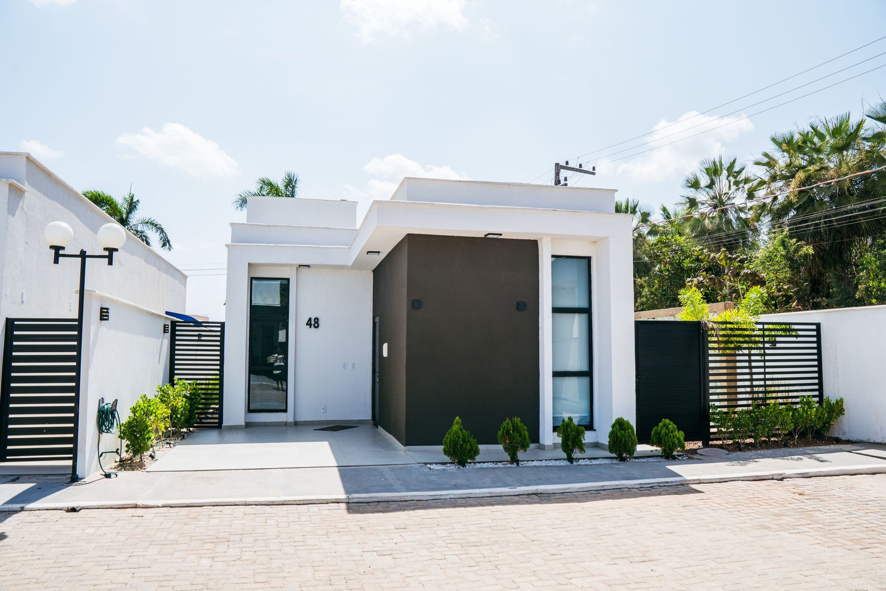 A modern two-story house with a flat roof, a large garage door, and a small front yard with potted plants.