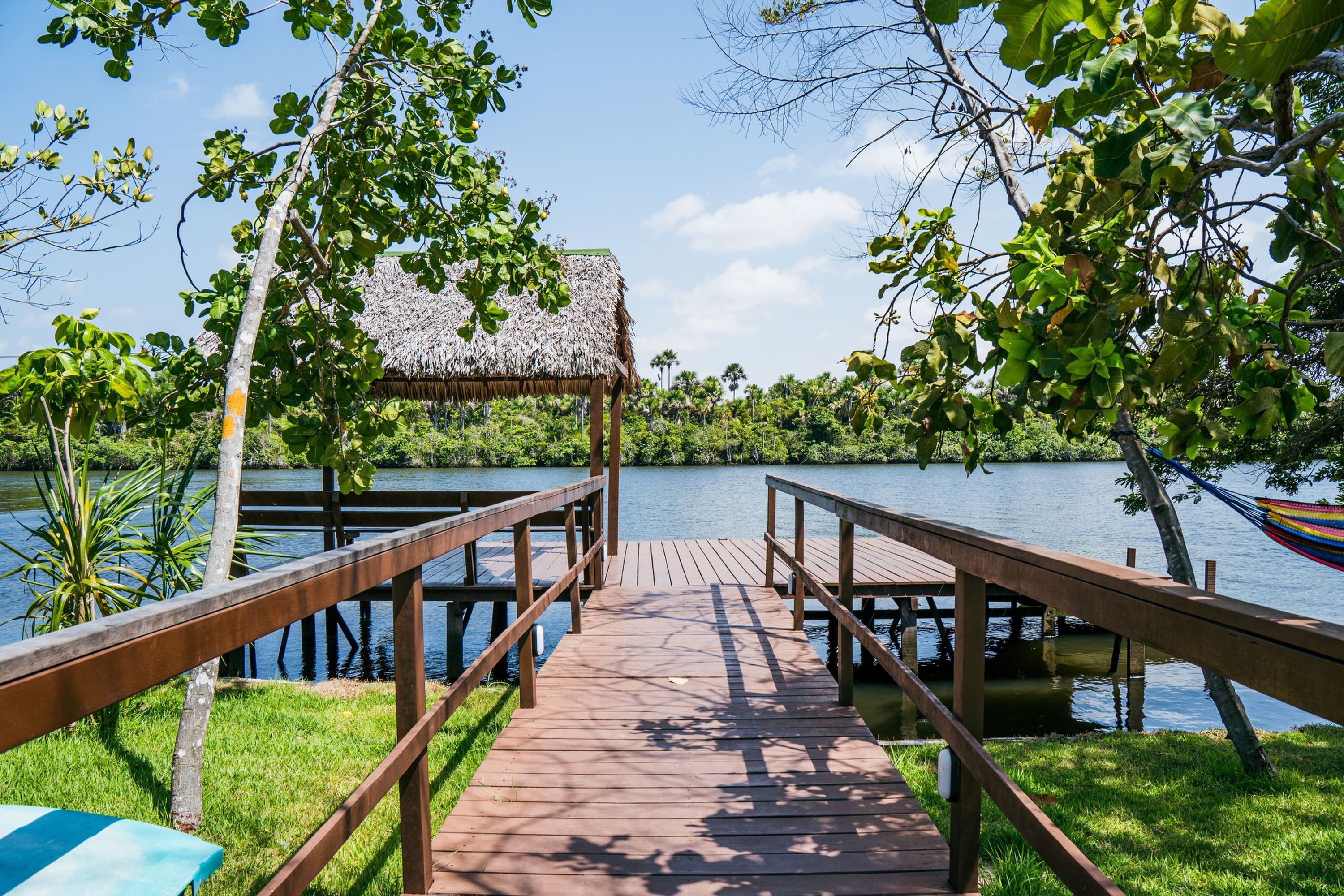 A wooden dock leading to a thatched-roof structure by a calm lake surrounded by lush greenery.