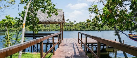 A wooden dock leading to a thatched-roof structure by a calm lake surrounded by lush greenery.