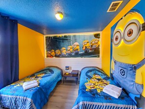 A room with two beds, a window with a Minion-themed artwork, a desk with a lamp, and a Minion figure.