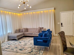 Ein Wohnzimmer mit einem Ecksofa, einem blauen Sessel, einem gemusterten Teppich und einer Deckenleuchte.