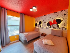 A hotel room with two beds, a window with curtains, a Mickey Mouse themed wall, and a ceiling light.