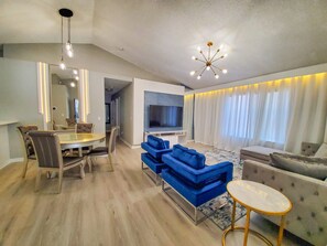 A modern living room with a dining area, a blue sofa set, a TV, and a chandelier.