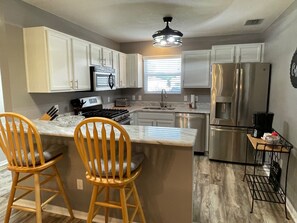 Una cocina con armarios blancos, un refrigerador de acero inoxidable, una estufa y dos taburetes de bar de madera.