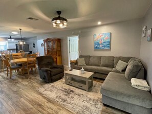 A living room with a grey sofa, a coffee table, a dining area with wooden chairs, and a painting on the wall.