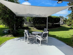Un patio al aire libre con mesa y sillas bajo una vela de sombra, rodeado de follaje.