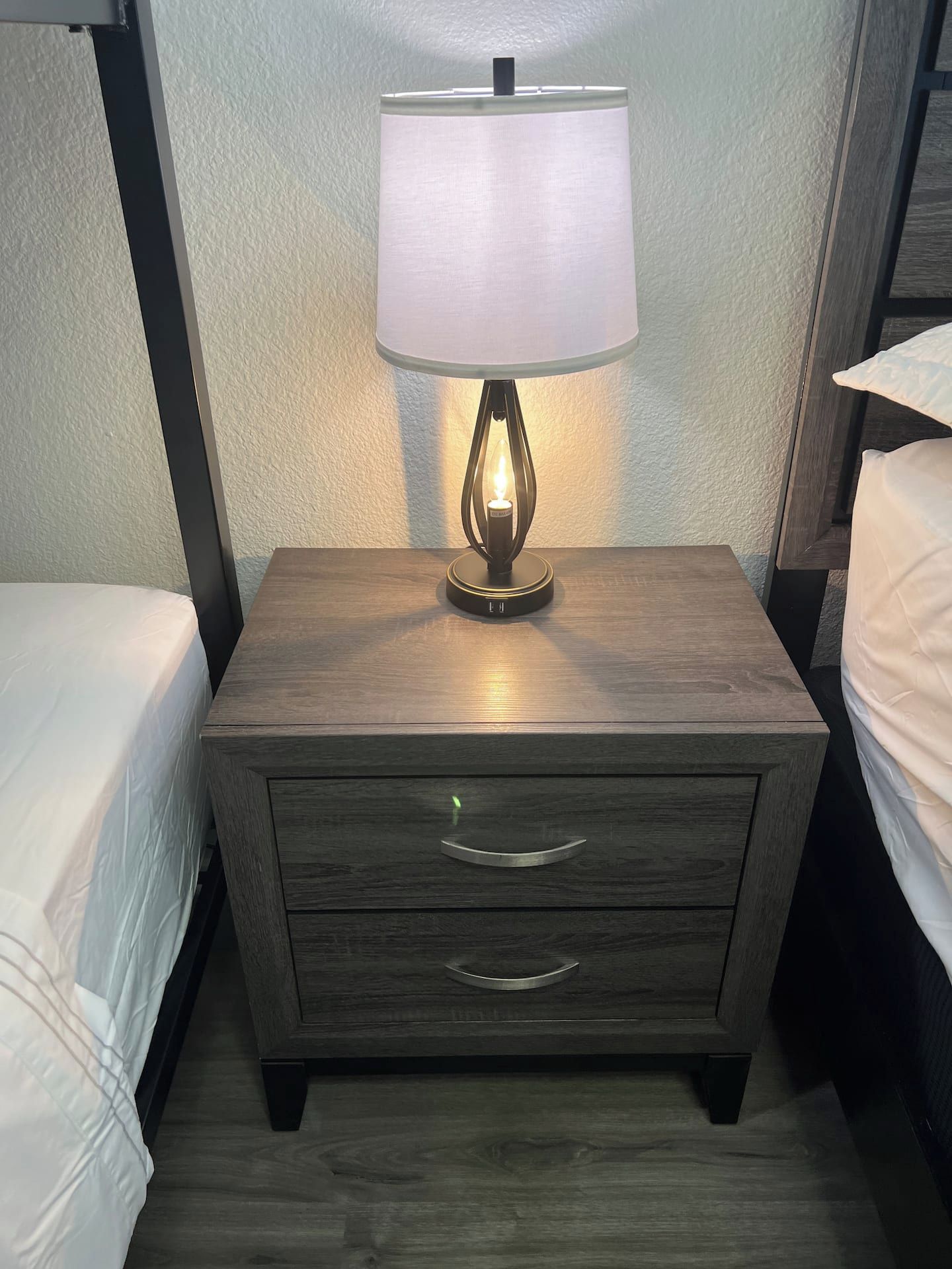 Uma mesa de cabeceira de madeira com uma luminária, duas gavetas e uma cama com roupa de cama branca.