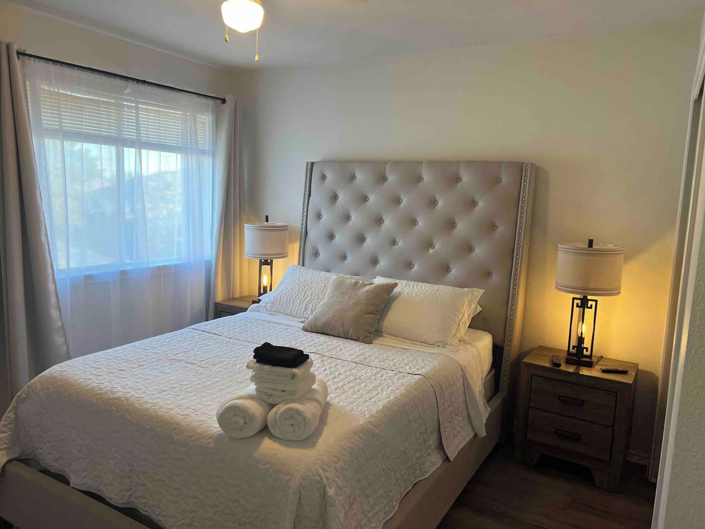 Una cama bien tendida con un cabecero capitonado, dos lámparas de noche y una ventana con cortinas.