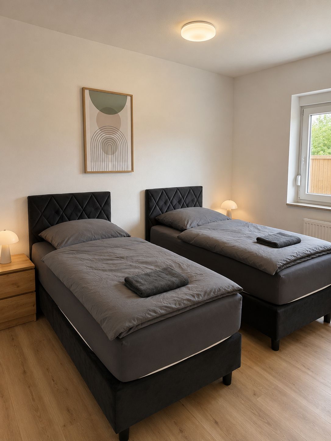 A bedroom with two beds, a bedside table, a lamp, a framed picture, and a window with a view.