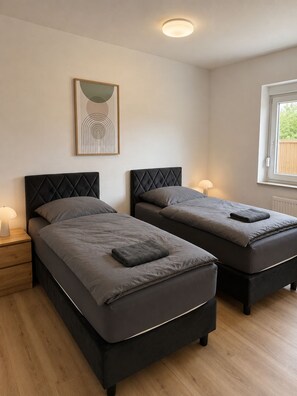 A bedroom with two beds, a bedside table, a lamp, a framed picture, and a window with a view. - Mühlhausen Ground Floor Apartment for Workers and Vacationers (Mühlhausen)