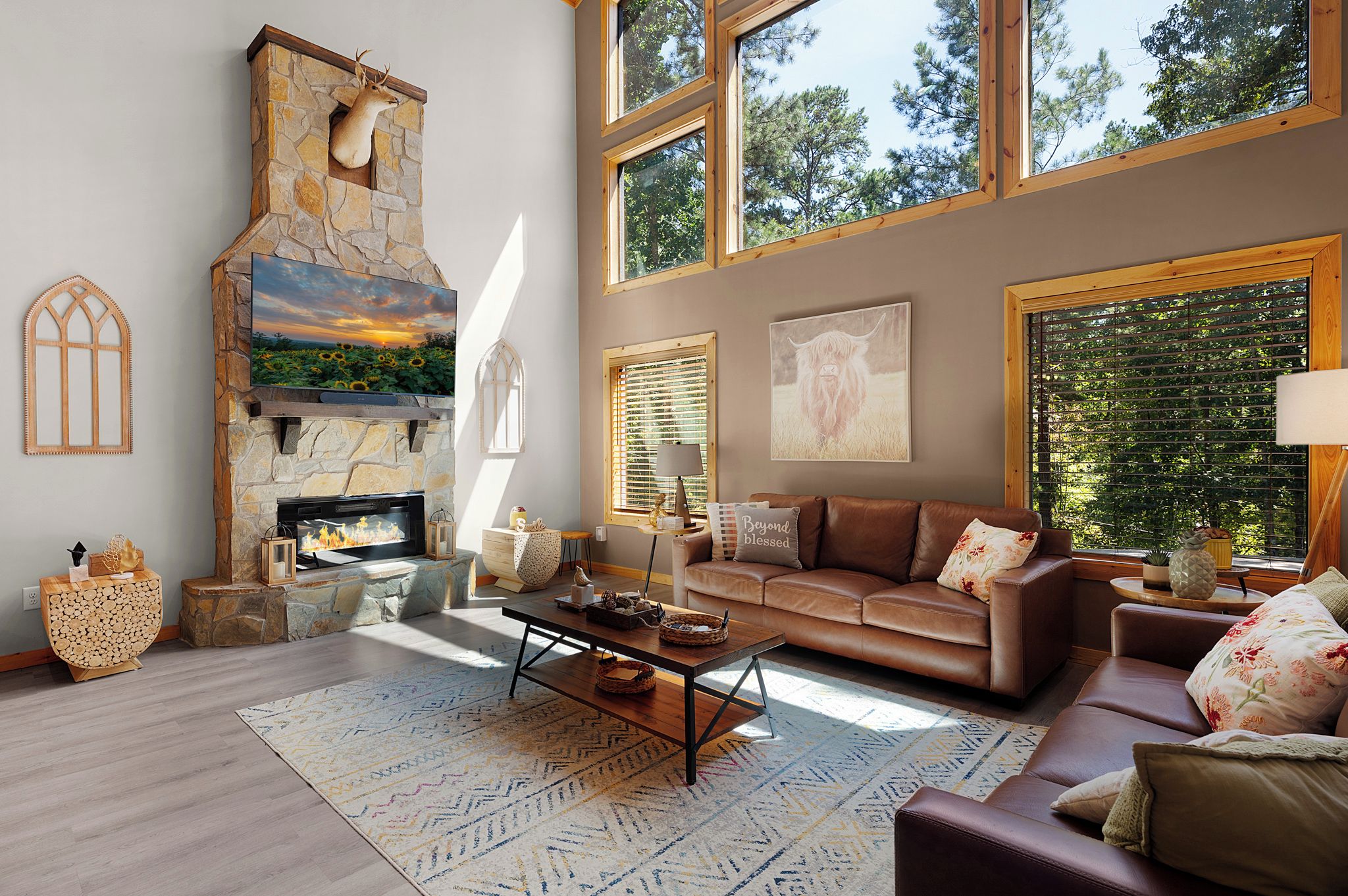 A spacious living room with a stone fireplace, a large window with blinds, a leather sofa, and a flat-screen TV mounted above the fireplace.