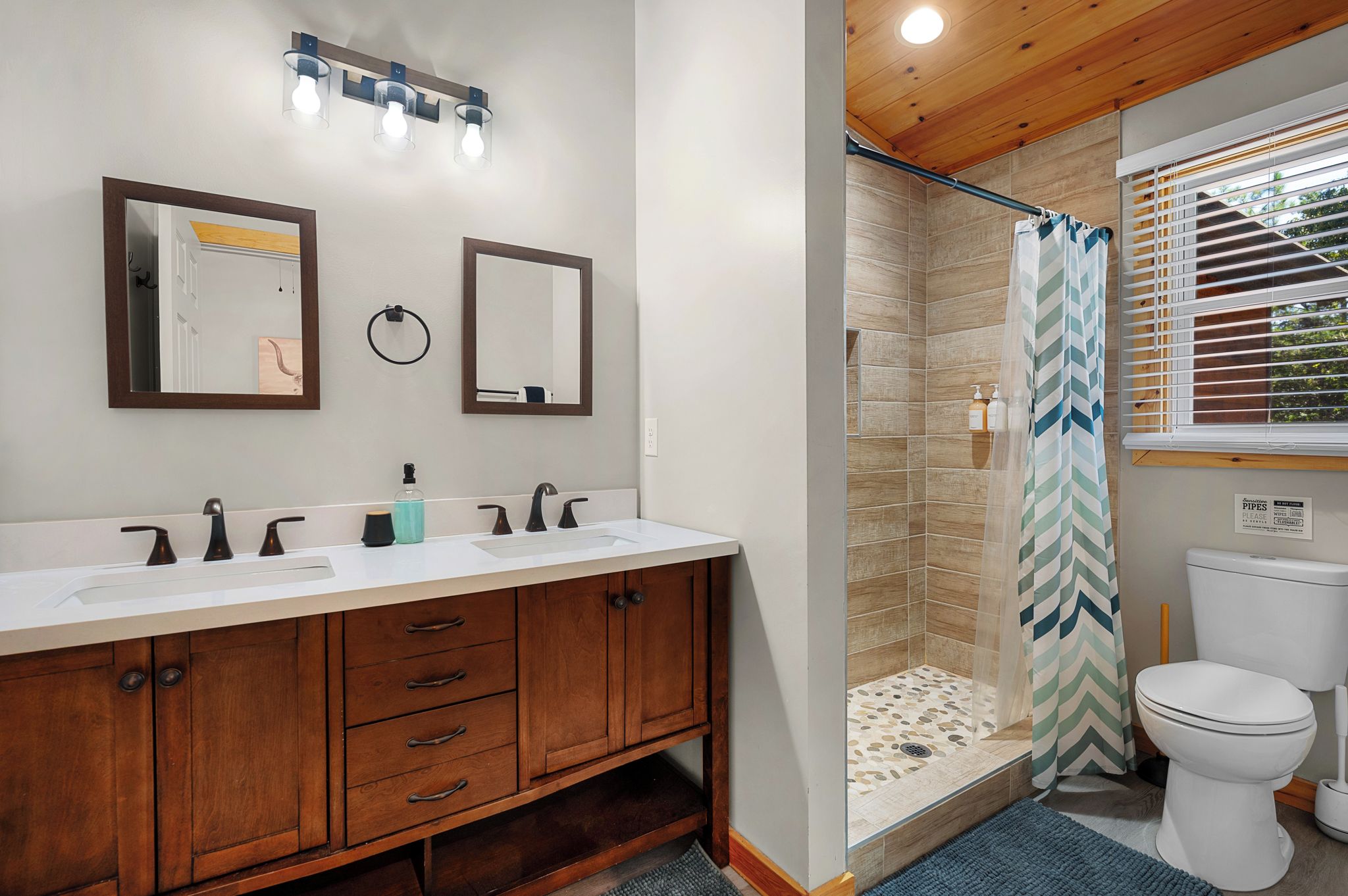 Un baño con tocador de madera, dos espejos, ducha con cortina a cuadros y un inodoro.