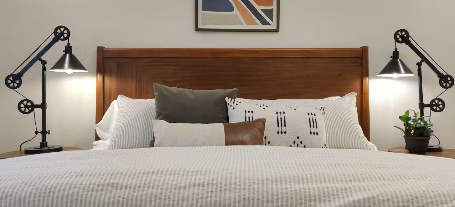 Un lit bien fait, agrémenté d’un coussin à motifs, d’un coussin marron et d’un coussin blanc. Deux lampes de bureau noires sont placées de chaque côté du lit.