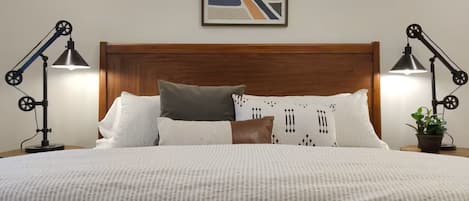 Un lit bien fait, agrémenté d’un coussin à motifs, d’un coussin marron et d’un coussin blanc. Deux lampes de bureau noires sont placées de chaque côté du lit.
