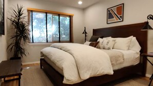 A bedroom with a bed, a nightstand, a lamp, a window with blinds, and a painting on the wall. - WFH Retreat at Jackson Market (Culver City)