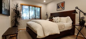Un dormitorio con cama, mesita de noche, lámpara, ventana con persianas y un cuadro en la pared.