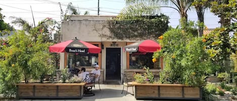 Un restaurante con mesas al aire libre, sombrillas rojas y una jardinera de madera con flores.