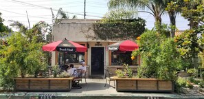 Un restaurante con mesas al aire libre, sombrillas rojas y una jardinera de madera con flores.