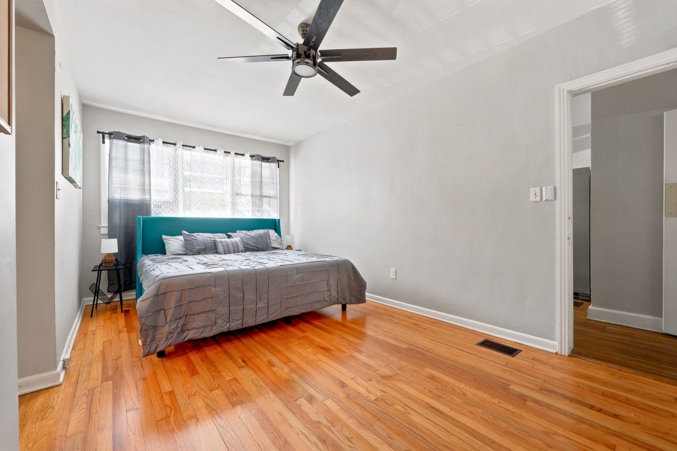 A bedroom with a bed, a ceiling fan, a window with curtains, and a wooden floor.