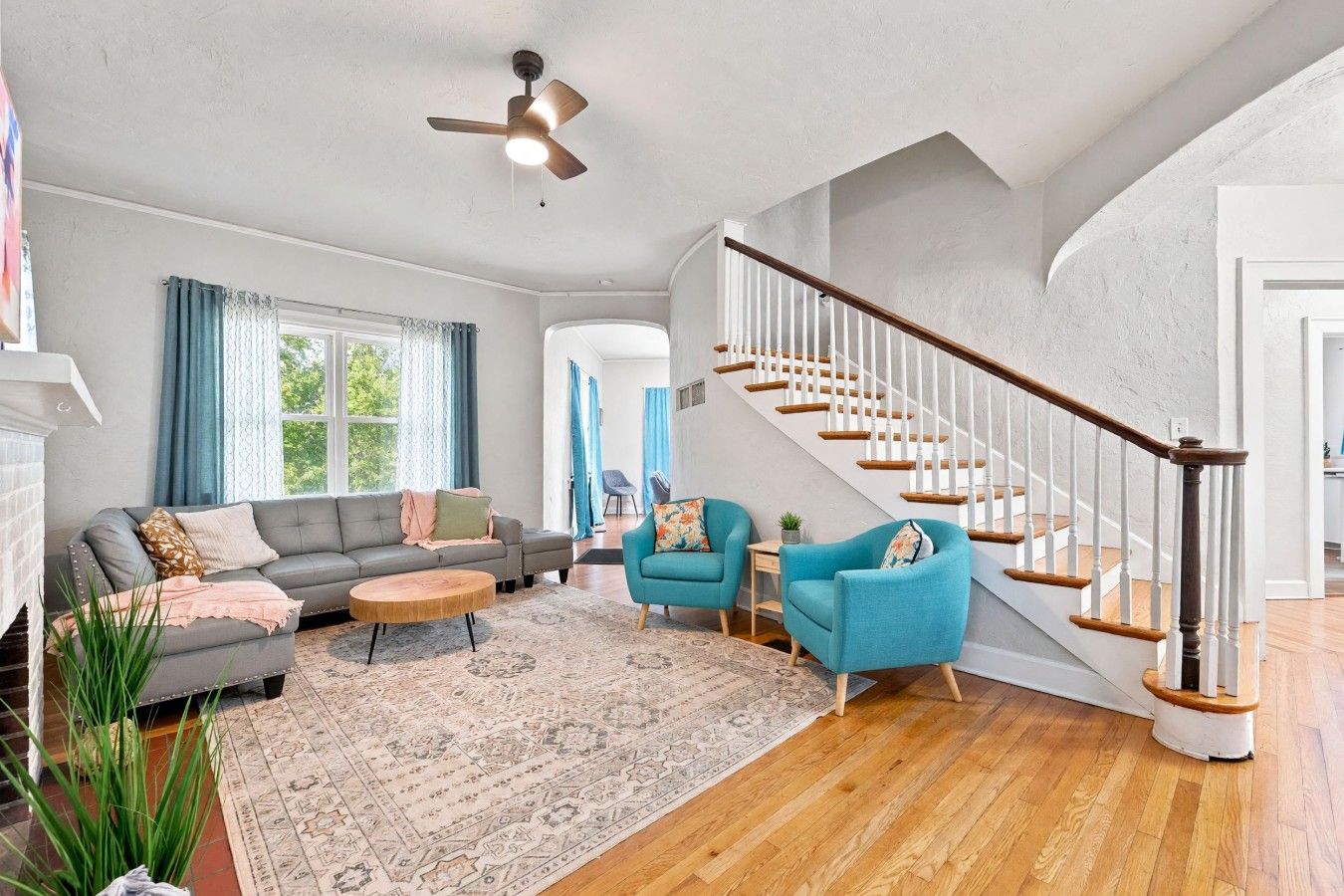 A spacious living room with a grey sofa, two blue armchairs, a wooden coffee table, and a patterned rug. A wooden staircase with white railings leads to another room.