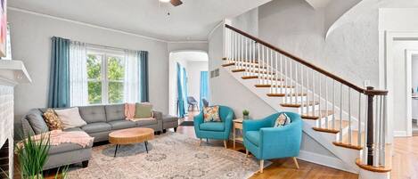 A spacious living room with a grey sofa, two blue armchairs, a wooden coffee table, and a patterned rug. A wooden staircase with white railings leads to another room.