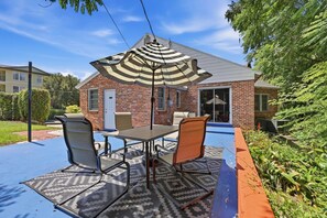 A patio with a table and chairs, a striped umbrella, and a brick house in the background. - Walk to UF  Shands Ultimate Tailgate House (Gainesville)