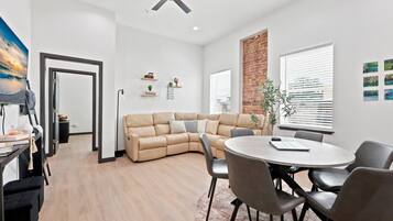 A modern living room with a beige sectional sofa, a round dining table with chairs, a ceiling fan, and a brick accent wall.