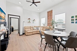 Una sala moderna con un sofá seccional color beige, una mesa redonda de comedor con sillas, un ventilador de techo y una pared decorativa de ladrillo.