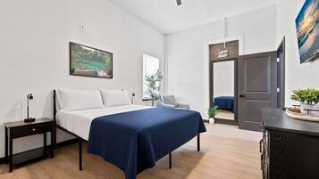A bedroom with a bed, bedside table, and a nightstand. There is a window with a view of greenery and a door leading to another room.
