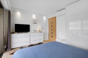 Un dormitorio moderno con una televisión de pantalla plana, una mesita de noche con lámpara, un armario y una cama con cubrecama azul.