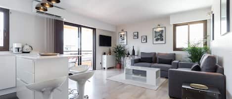 Una sala de estar moderna con un sofá gris, una mesa de centro y una zona de cocina con hervidor y cafetera.