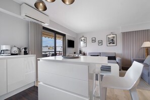 Una cocina moderna con armarios blancos, un comedor con mesa y sillas blancas, un sofá gris y un televisor montado en la pared.