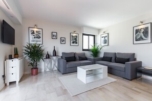 En moderne stue med en grå sofa, et sofabord og et fjernsyn ophængt på væggen. Flere billeder hænger på væggen, og der står en potteplante på gulvet.