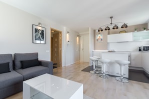 Un salon moderne avec un canapé gris, une table basse blanche et un coin cuisine doté de tabourets de bar blancs.