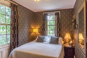 Une chambre à coucher avec un lit, deux tables de chevet, une fenêtre avec des rideaux et un mur recouvert d’un papier peint à motifs.