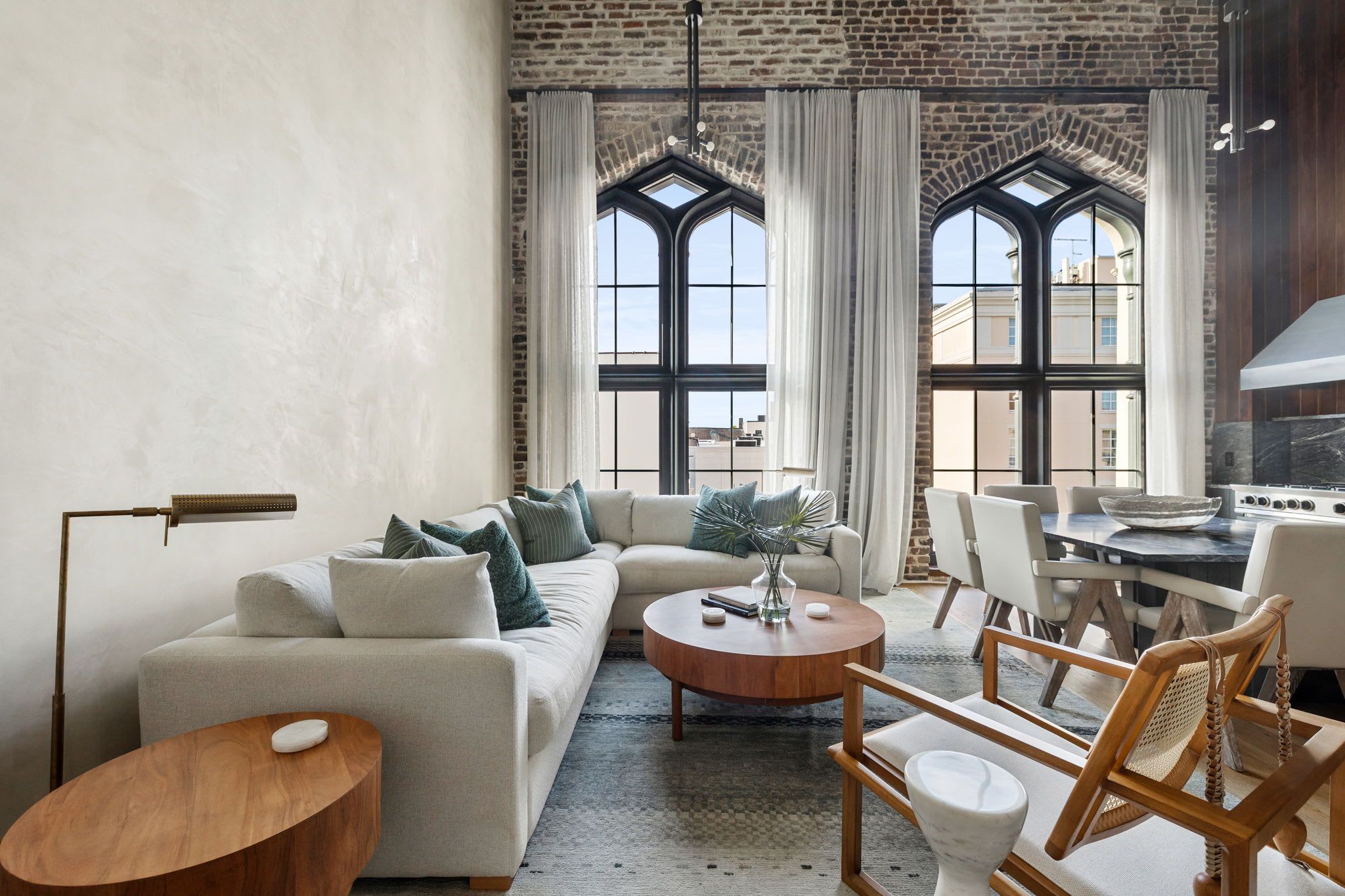 A modern living room with a large sofa, a coffee table, and a dining area with chairs. There are large windows with curtains, a fireplace, and a brick wall.