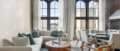 Un salon moderne avec un canapé d’angle, une table basse ronde et un coin repas avec des chaises. De grandes fenêtres aux montures noires et aux rideaux blancs sont visibles.