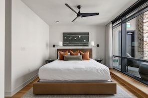 A modern bedroom with a large bed, a ceiling fan, a view of the outdoors, and a brick wall.